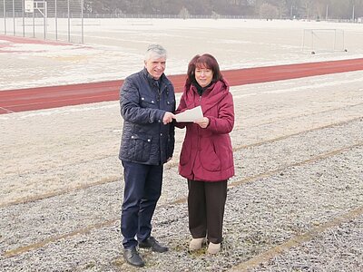 Zwei Personen stehen auf einem frostigen Sportplatz. Sie halten ein Dokument in der Hand und scheinen sich über etwas zu unterhalten. Im Hintergrund sind ein Laufbahn und ein Fußballfeld zu sehen.