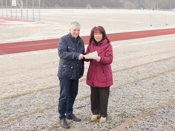 Zwei Personen stehen auf einem frostigen Sportplatz. Sie halten ein Dokument in der Hand und scheinen sich über etwas zu unterhalten. Im Hintergrund sind ein Laufbahn und ein Fußballfeld zu sehen.