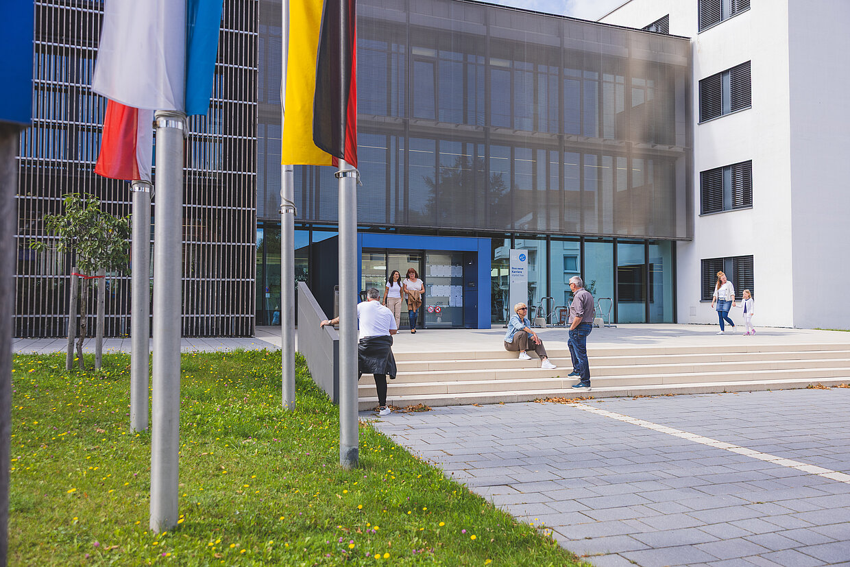 Eine moderne Gebäudeansicht mit einer Glasfassade. Im Vordergrund sind mehrere Personen zu sehen, die die Treppen hinauf- und hinuntergehen. Flaggen verschiedener Länder stehen links im Bild. Das Umfeld ist grün und freundlich gestaltet.