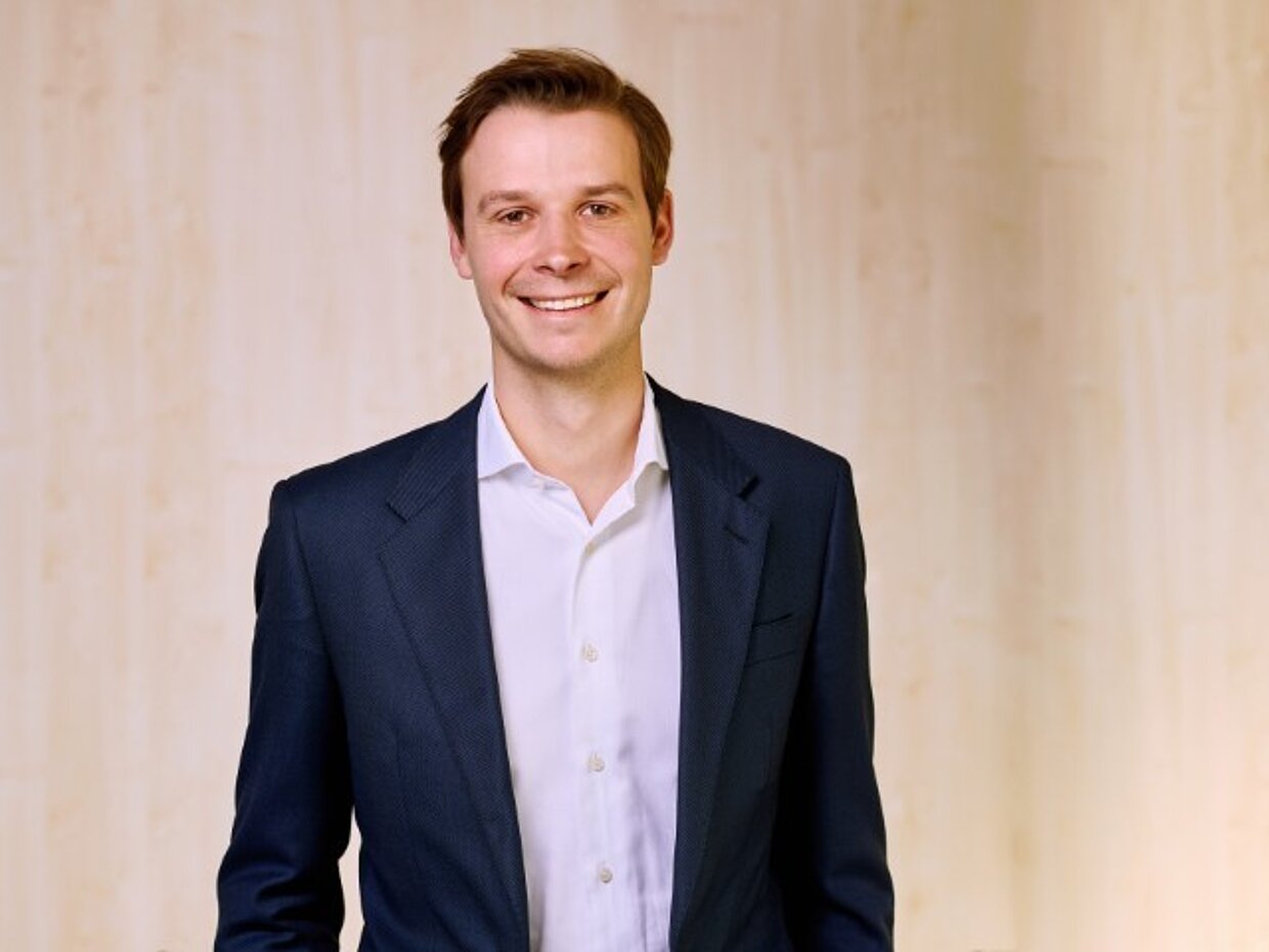 Ein lächelnder Mann in einem dunklen Anzug steht vor einem hellen Hintergrund. Er trägt ein weißes Hemd und hat kurzes, unordentliches Haar. Die Pose ist freundlich und selbstbewusst.