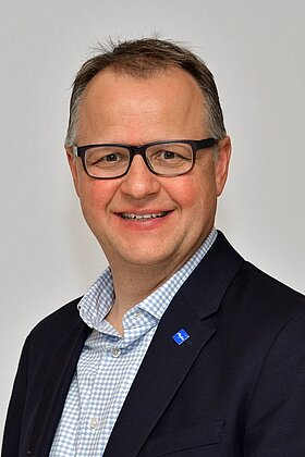 Porträt eines lächelnden Mannes mit Brille, der einen dunkelblauen Blazer und ein kariertes Hemd trägt. Er steht vor einem neutralen Hintergrund und schaut freundlich in die Kamera.
