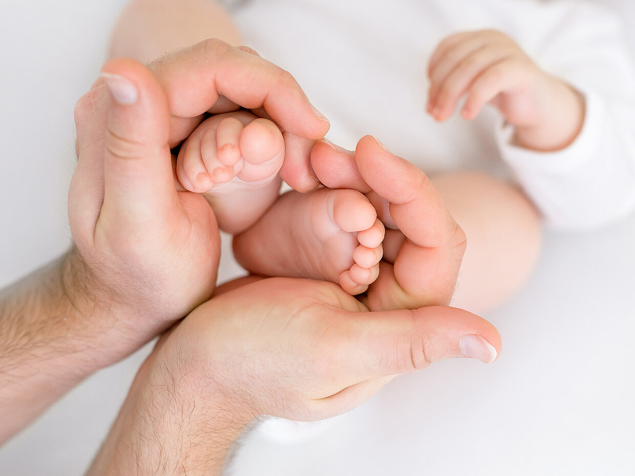 Ein Erwachsener hält die Füße eines Babys in seinen Händen. Die Füße sind klein und zart, während die Hände den Schutz und die Fürsorge symbolisieren. Im Hintergrund sieht man einen Teil des Babys, das in einem weißen Outfit gekleidet ist.