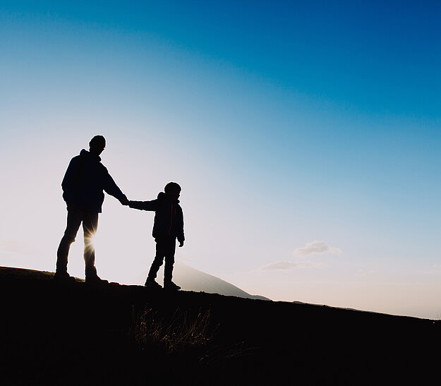 Silhouette eines Erwachsenen und eines Kindes, die Hand in Hand bei Sonnenuntergang stehen. Der Himmel zeigt sanfte Blau- und Orangetöne. Die Umgebung ist unkenntlich, während sich die Figuren deutlich abheben.
