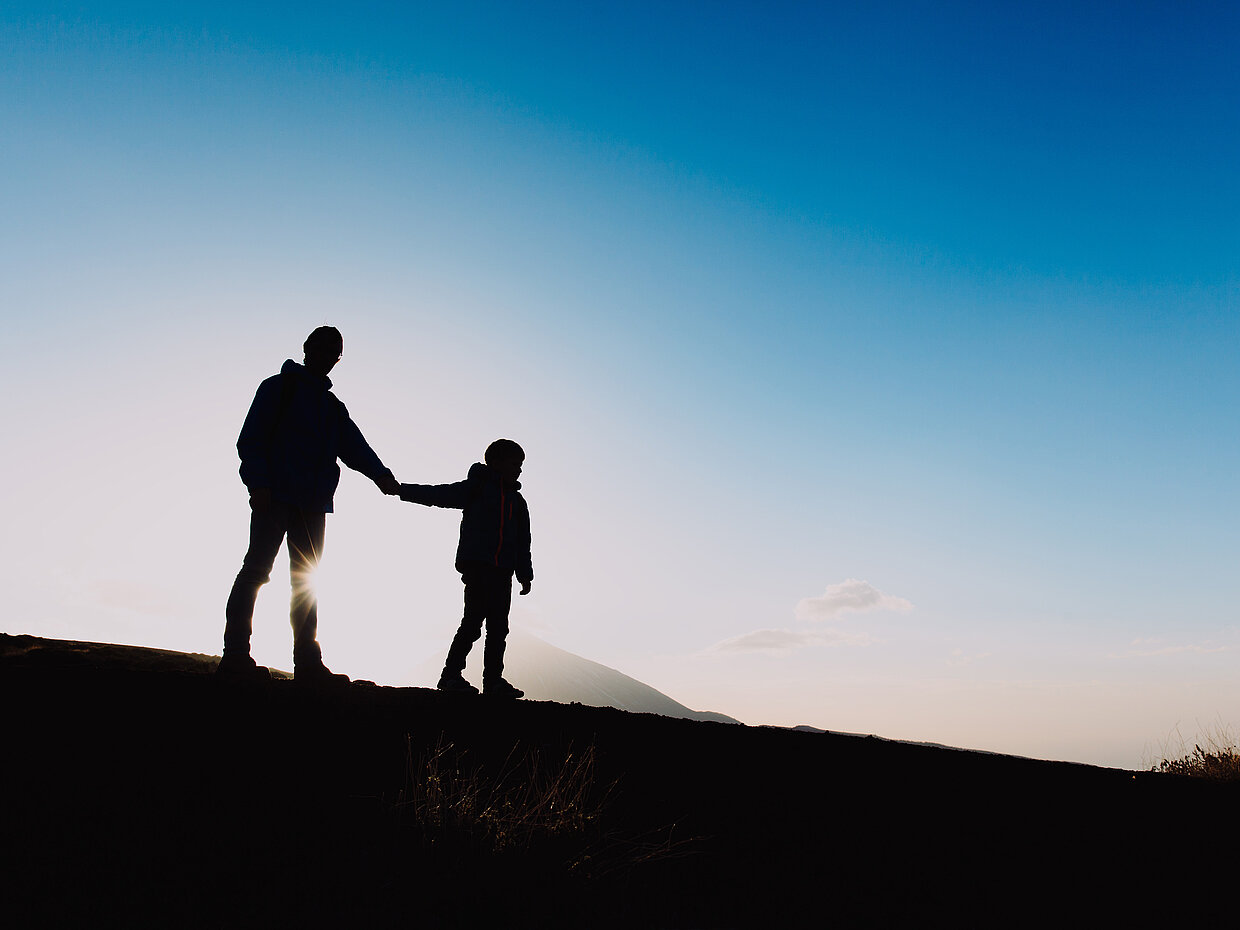 Silhouette eines Erwachsenen und eines Kindes, die Hand in Hand bei Sonnenuntergang stehen. Der Himmel zeigt sanfte Blau- und Orangetöne. Die Umgebung ist unkenntlich, während sich die Figuren deutlich abheben.
