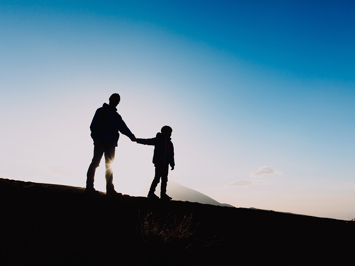Silhouette eines Erwachsenen und eines Kindes, die Hand in Hand bei Sonnenuntergang stehen. Der Himmel zeigt sanfte Blau- und Orangetöne. Die Umgebung ist unkenntlich, während sich die Figuren deutlich abheben.