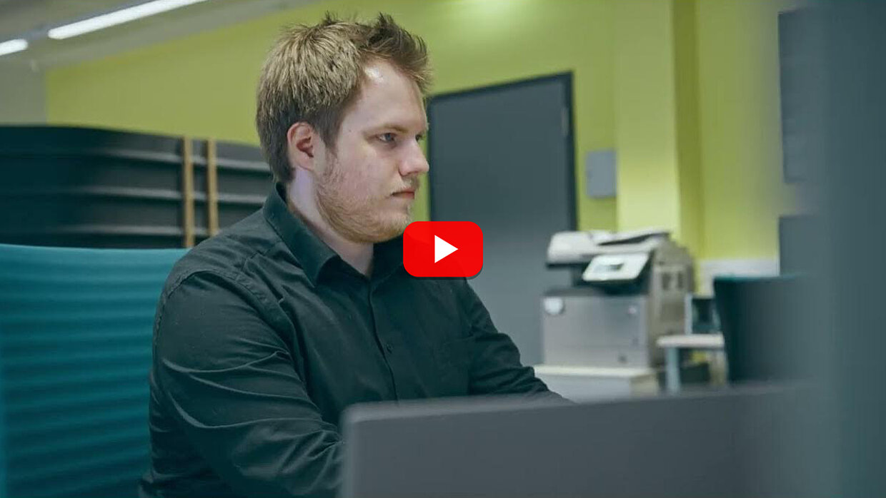 Zum Video: Ausbildung beim Landratsamt Ostallgäu (YouTube) Ein Mann mit kurzen, blonden Haaren sitzt an einem Schreibtisch und schaut konzentriert auf einen Laptop. Im Hintergrund sind Regale und ein Drucker sichtbar. Die Wand ist hellgrün gestrichen. Ein YouTube-Play-Button ist in der Mitte des Bildes platziert.