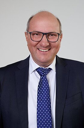 Ein lächelnder Mann mit Brille, der einen Anzug und eine Krawatte trägt. Er steht vor einer hellen Wand und hat eine freundliche Ausstrahlung.