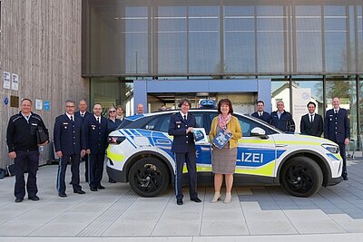 Eine Gruppe von Polizeibeamten und -beamten steht vor einem neuen Polizeiauto. Sie posieren lächelnd vor einem modernen Gebäude. Einige Personen tragen Uniformen, und eine Frau in ziviler Kleidung hält eine Broschüre. Das Auto ist mit auffälligen Farben markiert.