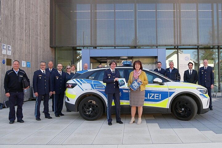 Eine Gruppe von Polizeibeamten und -beamten steht vor einem neuen Polizeiauto. Sie posieren lächelnd vor einem modernen Gebäude. Einige Personen tragen Uniformen, und eine Frau in ziviler Kleidung hält eine Broschüre. Das Auto ist mit auffälligen Farben markiert.