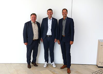 Drei Männer in Anzügen stehen neben einem weißen Hintergrund. Der Mann links trägt ein helles Hemd, während die anderen zwei dunkelblau gekleidete Anzüge tragen. Alle lächeln in die Kamera.