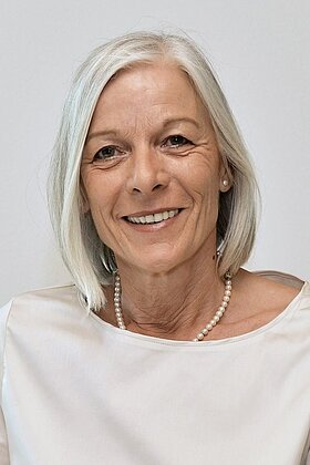 Porträt einer lächelnden Frau mit schulterlangem, grauem Haar. Sie trägt eine elegante, helle Bluse und eine Perlenkette. Der Hintergrund ist neutral, umfokussiert auf ihren freundlichen Gesichtsausdruck.