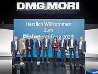 Eine Gruppe von zehn Personen steht auf einer Bühne vor einem großen Banner mit der Aufschrift „DMG MORI“ und „Herzlich Willkommen Zum Dialogeröffnung 2023“. Die Personen sind geschäftlich gekleidet und lächeln zur Kamera.