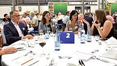 Eine Gruppe von vier Personen sitzt an einem runden Tisch bei einem Event. Sie unterhalten sich lebhaft, während auf dem Tisch Gläser mit Wasser und Rotwein sowie Geschirr sichtbar sind. Im Hintergrund sind weitere Gäste und ein Banner zu sehen.