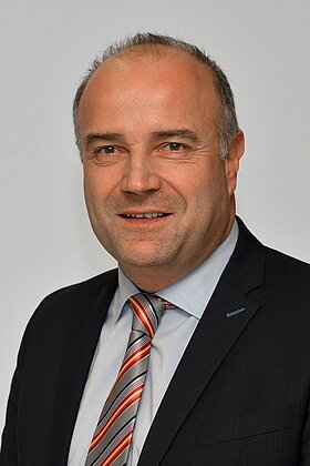 Porträt eines Mannes in Anzug mit Krawatte. Er hat kurzes, graues Haar und lächelt freundlich. Der Hintergrund ist hell und neutral.