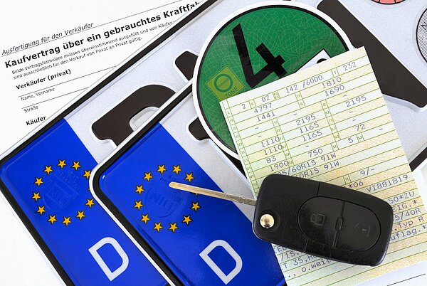 Ein Kfz-Kennzeichen mit europäischen Symbolen, ein Fahrzeugschlüssel, ein Kaufvertrag für ein gebrauchtes Kraftfahrzeug und eine Prüfplakette liegen auf einem Tisch. Neben diesen Dokumenten sind weitere Papierrechnungen sichtbar.