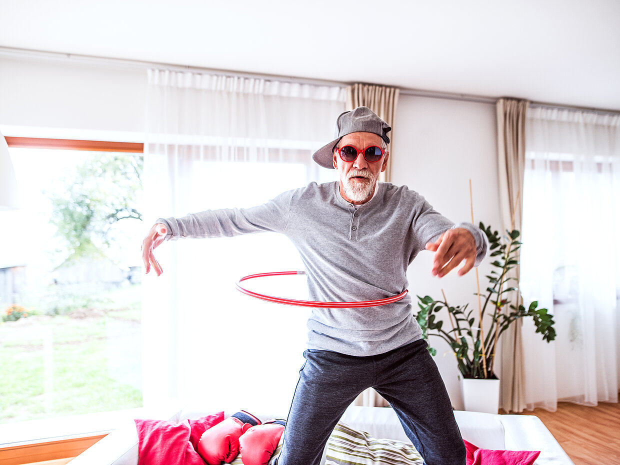 Ein älterer Mann mit einer Sonnenbrille und einer Mütze steht in einem hellen Raum und dreht einen Hula-Hoop-Reifen um seine Taille. Er trägt ein graues Oberteil und dunkle Hose. Im Hintergrund sind Fenster mit Tageslicht und Pflanzen zu sehen.