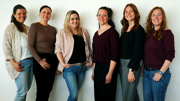 Sechs Frauen stehen in einer Reihe vor einer weißen Wand. Sie tragen unterschiedliche Kleidung und lächeln. Die Frauen haben verschiedene Haarfarben und -längen. Die Stimmung ist freundlich und entspannt.