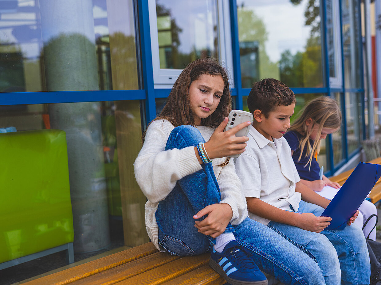 Drei Kinder sitzen auf einer Bank vor einem Glasgebäude. Ein Mädchen hält ein Smartphone, während ein Junge daneben auf ein Blatt Papier schaut. Ein weiteres Mädchen beschäftigt sich mit einem Laptop. Alle wirken konzentriert und nachdenklich.