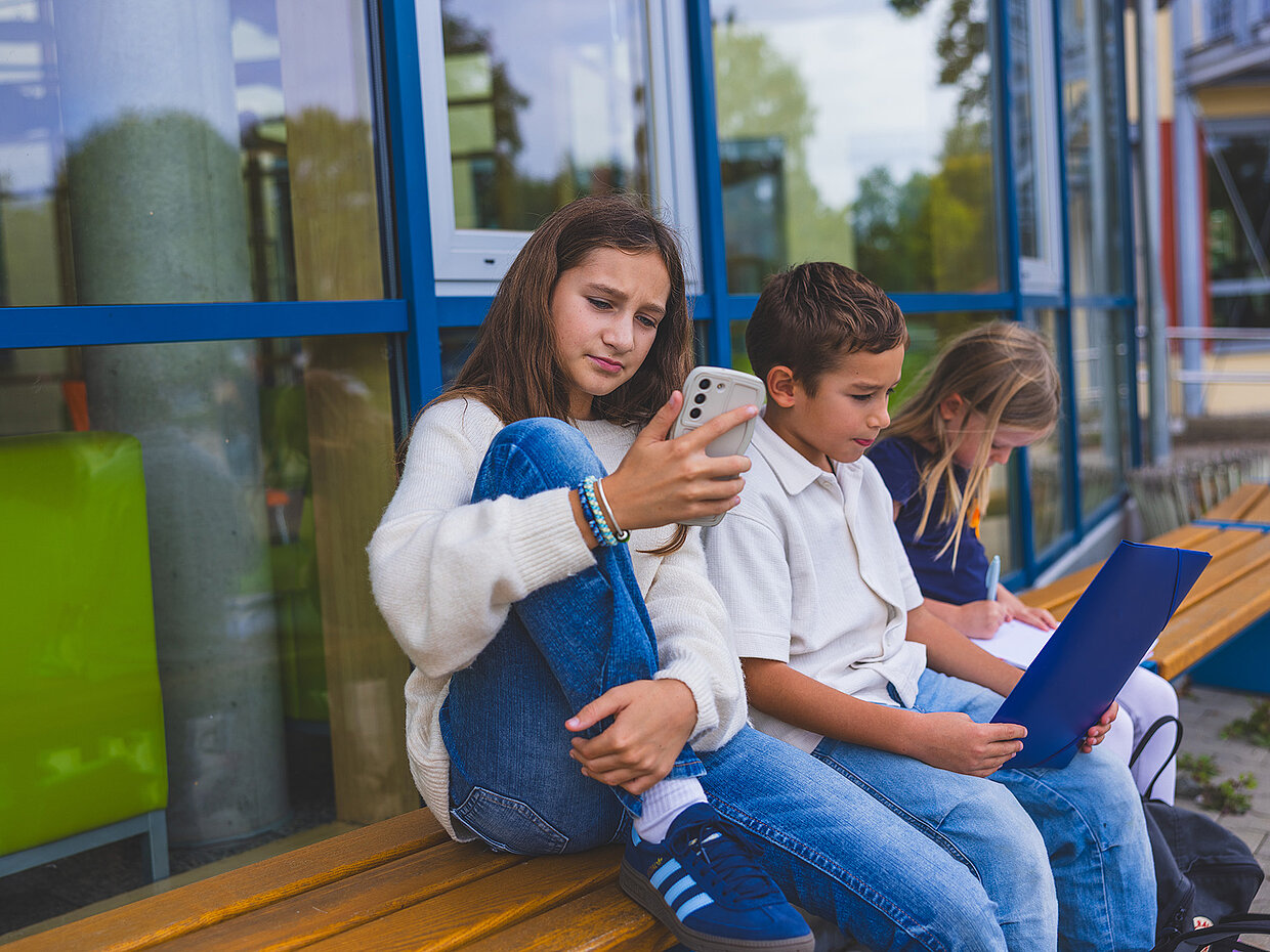 Drei Kinder sitzen auf einer Bank vor einem Glasgebäude. Ein Mädchen hält ein Smartphone, während ein Junge daneben auf ein Blatt Papier schaut. Ein weiteres Mädchen beschäftigt sich mit einem Laptop. Alle wirken konzentriert und nachdenklich.