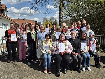 Eine Gruppe von Menschen steht auf einer Terrasse und hält Urkunden in den Händen. Sie lächeln in die Kamera. Im Hintergrund sind Bäume und ein blauer Himmel zu sehen. Einige Personen halten Blumen.