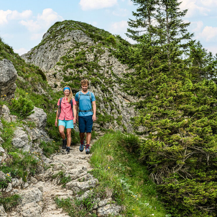 © Tourismusverband Ostallgäu e.V. / Peter von Felbert Zwei Wanderer auf einem schmalen Pfad in den Bergen, umgeben von Felsen und Bäumen. Im Hintergrund sind weitere Berge und ein bewölkter Himmel sichtbar. Ein Wegweiserschild steht in der Nähe.