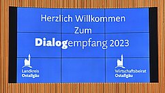 Altes Bild von einer digitalen Anzeige mit dem Text: "Herzlich Willkommen Zum Dialogempfang 2023". Die Hintergrundfarbe ist blau, mit Logos des Landkreises Ostallgäu und des Wirtschaftsbeirats Ostallgäu.