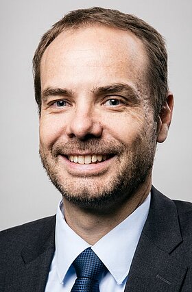 Porträt eines lächelnden Mannes mit kurzem, leicht gewelltem Haar und einem kurzen Bart. Er trägt einen dunklen Anzug mit einer hellblauen Hemd und einer blauen Krawatte. Der Hintergrund ist neutral und verschwommen.