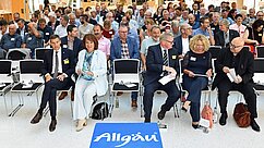Eine große Gruppe von Menschen sitzt in einem Konferenzraum mit Stühlen in mehreren Reihen. Im Vordergrund sitzen einige Personen, darunter eine Frau in einem hellen Anzug. Vor ihnen liegt ein Schild mit der Aufschrift „Allgäu“. Die Atmosphäre ist geschäftig und interessiert.