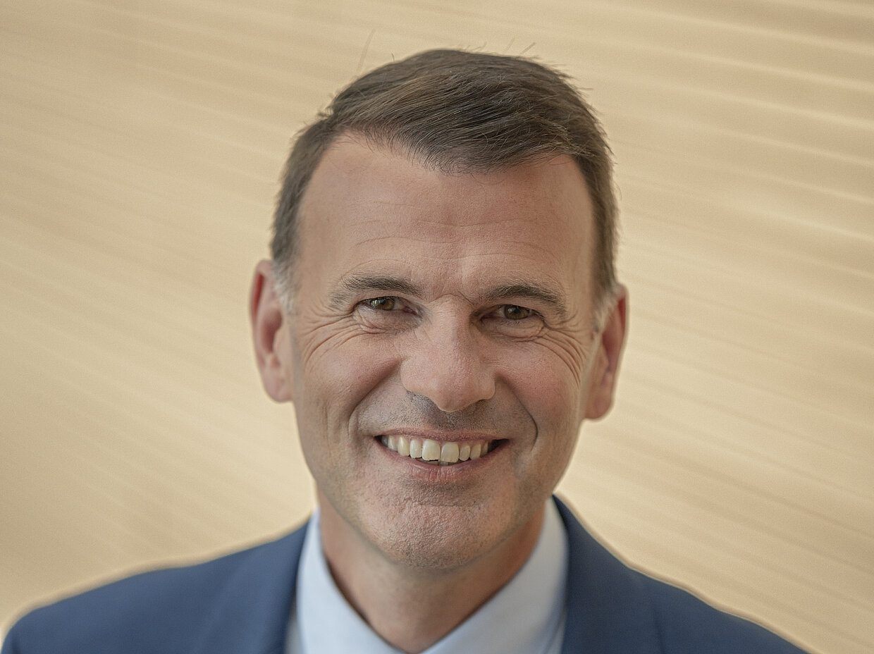 Ein gut gekleideter Mann mit kurzen, dunklen Haaren und einem freundlichen Lächeln steht vor einem neutralen, hellen Hintergrund. Er trägt einen hellblauen Anzug und eine passende Krawatte.