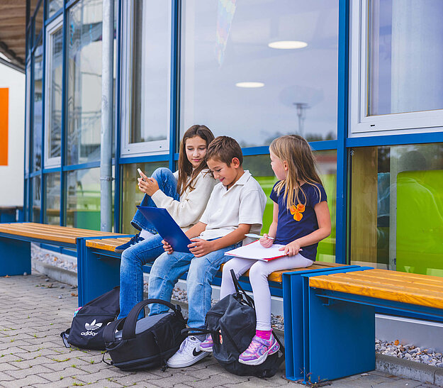 Das Bild zeigt 3 Schulkinder sitzend auf einer Bank vor dem Schulgebäude