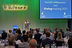 Eine Person steht an einem Rednerpult und begrüßt die Gäste eines Events mit einer Präsentation im Hintergrund. Tische sind mit weißen Tischtüchern und Gläsern gedeckt. Das Event findet in einem großen Raum statt, mit einer Bühnenbeleuchtung und einem Banner im Hintergrund.