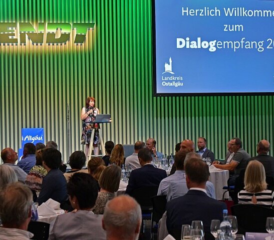 Eine Person steht an einem Rednerpult und begrüßt die Gäste eines Events mit einer Präsentation im Hintergrund. Tische sind mit weißen Tischtüchern und Gläsern gedeckt. Das Event findet in einem großen Raum statt, mit einer Bühnenbeleuchtung und einem Banner im Hintergrund.