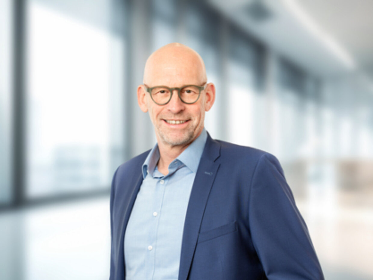 © Rösle GmbH & Co. KG Ein Mann mit Glatze und Brille lächelt in einem modernen Büro, das durch große Fenster lichtdurchflutet ist. Er trägt einen blauen Anzug und ein hellblaues Hemd. Der Hintergrund wirkt unauffällig und professionell.