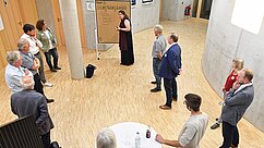 Eine Gruppe von Menschen steht in einem modernen Raum. Eine Frau präsentiert vor einem Plakat, während die anderen aufmerksam zuhören. Im Vordergrund sind Stehtische mit Getränken zu sehen. Der Raum hat helle Holzfußböden und Wände aus Beton.