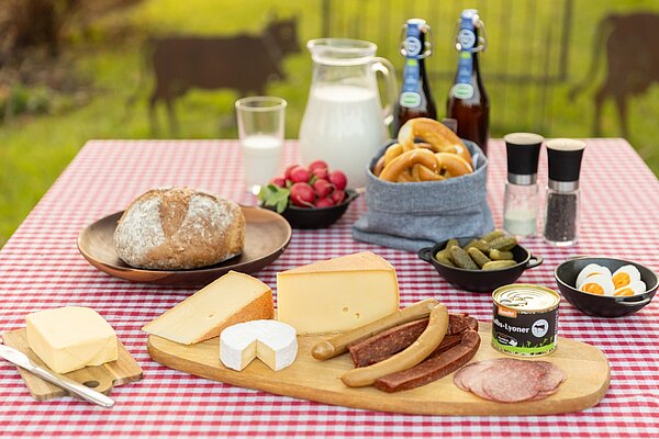 Ein gedeckter Tisch im Freien mit rot-weißem Karomuster. Auf einem Holzbrett sind verschiedene Käsesorten, Wurst, Butter und ein Becher Quark angerichtet. Daneben befinden sich Brot, Senf, eingelegte Gurken und Radieschen. Im Hintergrund stehen Milch und zwei Flaschen Bier.