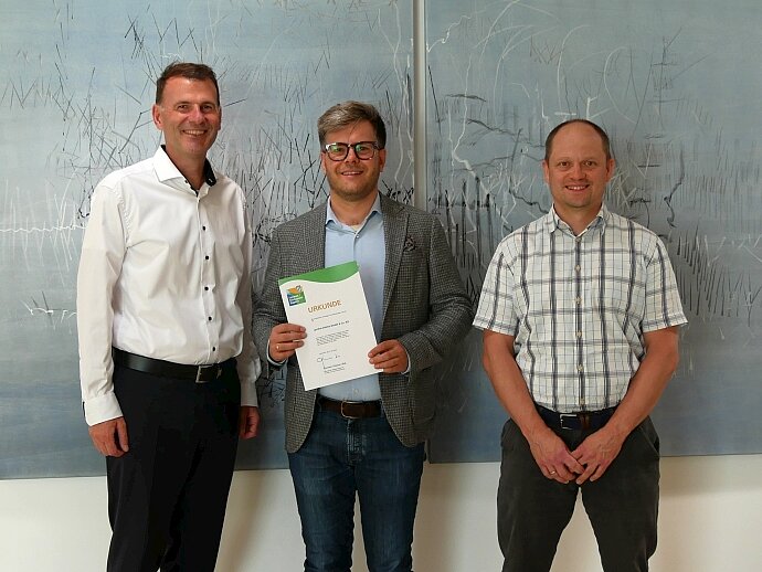 Drei Männer stehen nebeneinander vor einem abstrakten Kunstwerk. Der Mann in der Mitte hält ein Zertifikat in der Hand, während die anderen beiden ihm zugewandt sind. Sie tragen formelle Kleidung und stehen in einer hellen, modernen Umgebung.