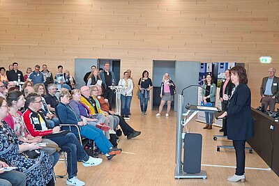 Eine Gruppe von Menschen sitzt und steht in einem Raum. Eine Rednerin steht an einem Pult und spricht zu dem Publikum. Die Zuhörer scheinen aufmerksam zuzuhören. Die Atmosphäre wirkt informell und einladend.