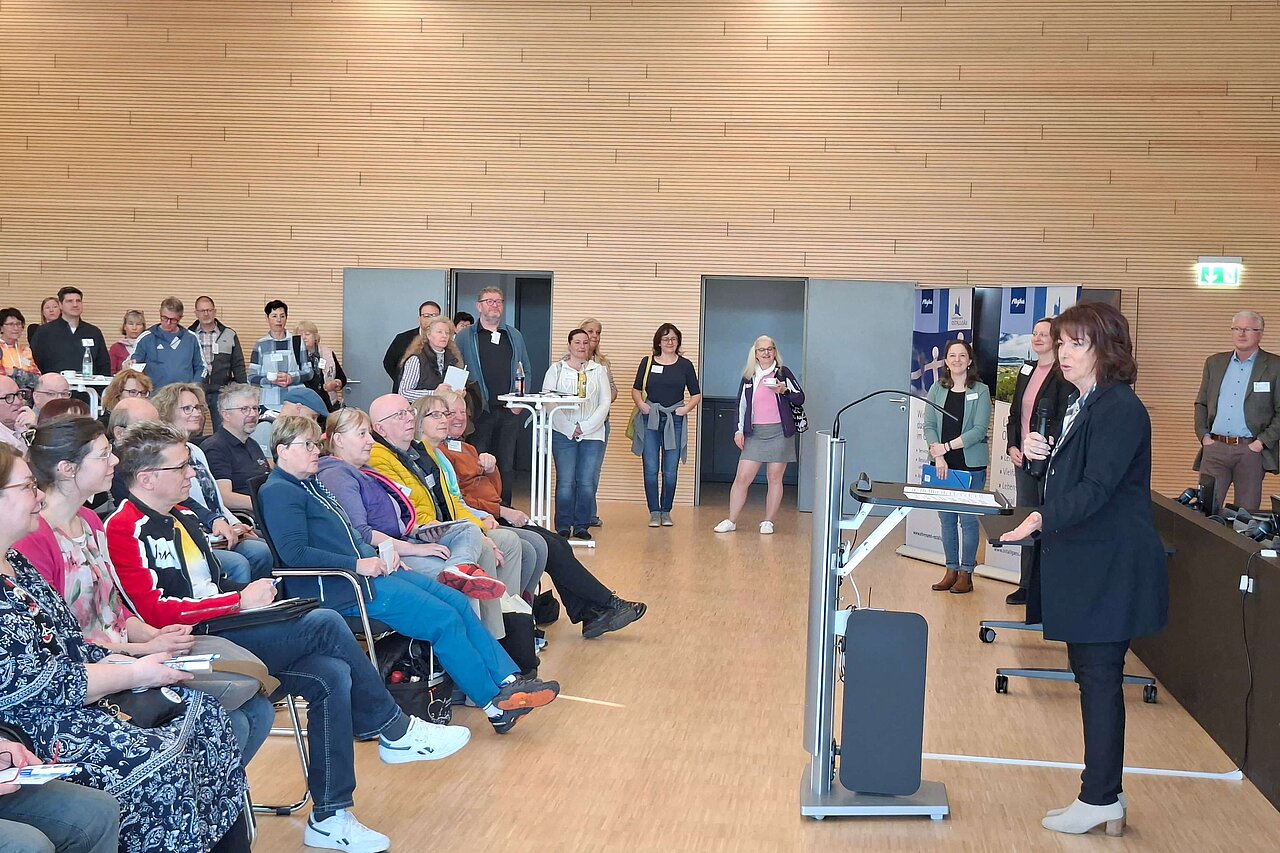 Eine Gruppe von Menschen sitzt und steht in einem Raum. Eine Rednerin steht an einem Pult und spricht zu dem Publikum. Die Zuhörer scheinen aufmerksam zuzuhören. Die Atmosphäre wirkt informell und einladend.