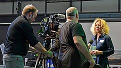 Ein Kamerateam interviewt eine Frau mit lockigem, blondem Haar. Der Mann links bedient die Kamera, während ein weiterer Mann rechts Mikrofone hält. Im Hintergrund sind verschwommene Bühnen- und Veranstaltungselemente zu erkennen.