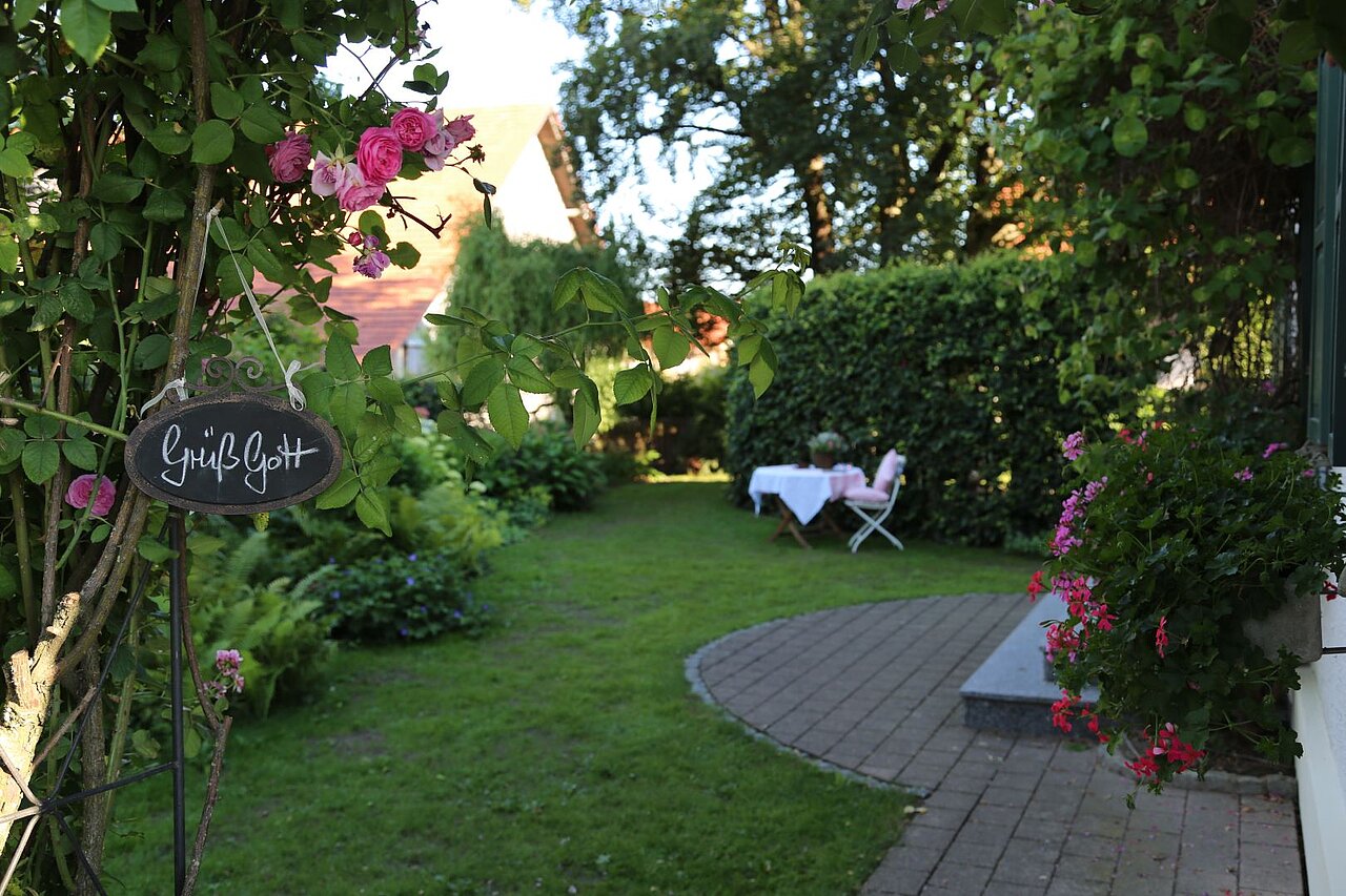 Ein schöner Garten mit grüner Rasenfläche und blühenden Pflanzen. Links hängt ein Schild mit der Aufschrift „Gott sei Dank“. Im Hintergrund steht ein Tisch mit weißen Tischdecken und zwei Stühlen, umgeben von Büschen und Bäumen.