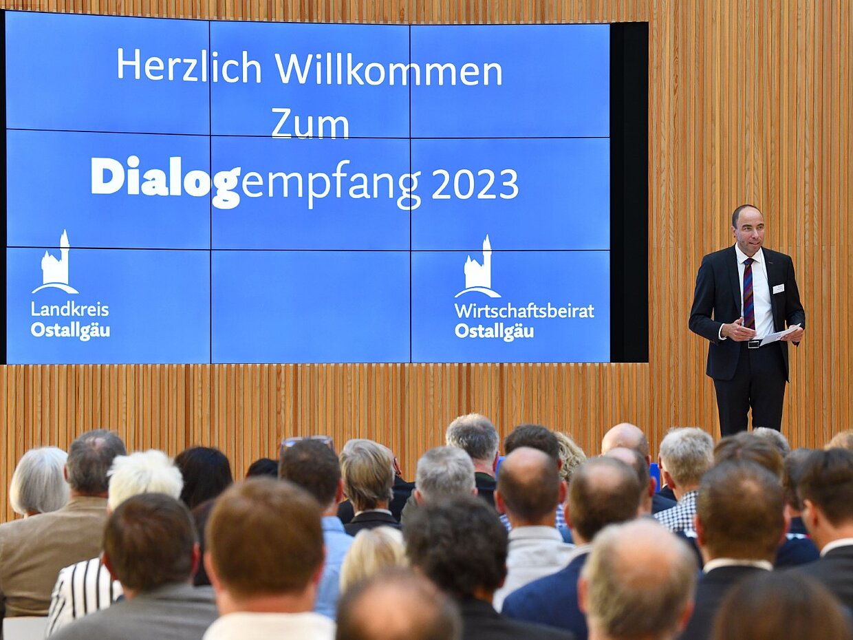 Ein Mann steht auf einer Bühne vor einer großen Anzeige, die für den "Dialogempfang 2023" im Landkreis Ostallgäu wirbt. Im Publikum sitzen viele Menschen, die interessiert zuhören. Die Szenerie vermittelt eine Atmosphäre des Dialogs und der Gemeinschaft.