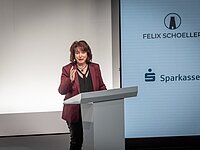 Eine Frau steht an einem Rednerpult und spricht. Sie trägt eine rot-schwarze Jacke und hat lange, braune Haare. Im Hintergrund sind die Logos von "Felix Schoeller" und "Sparkasse Altstadt" zu sehen. Die Szene scheint bei einer Veranstaltung oder Präsentation stattzufinden.