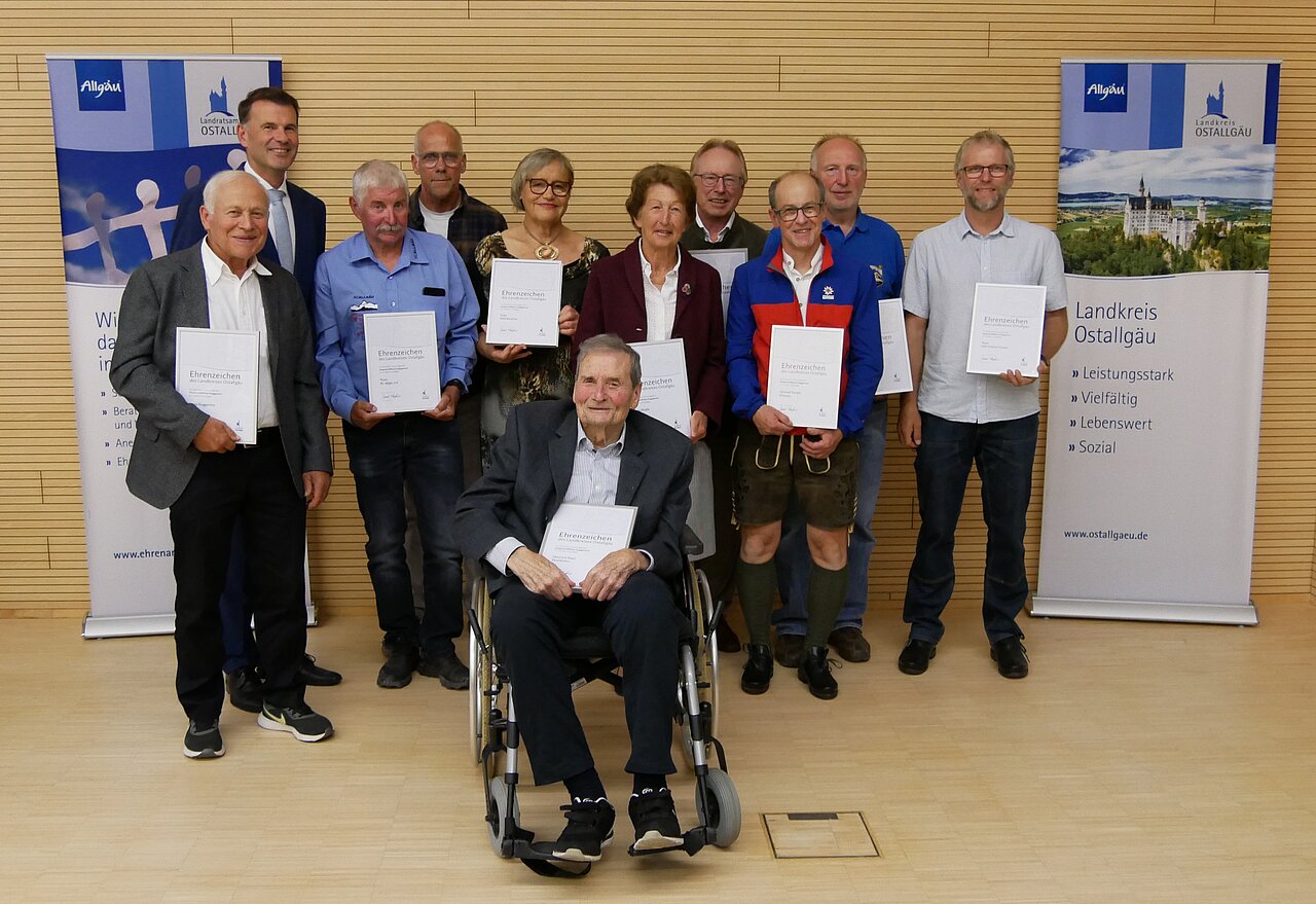 Eine Gruppe von neun Personen, darunter ein Mann im Rollstuhl, steht vor zwei Plakaten. Alle halten Urkunden in den Händen. Die Personen lächeln, es sind verschiedene Altersgruppen vertreten. Der Hintergrund ist hell und schlicht.