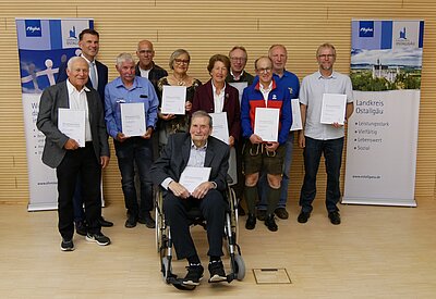 Eine Gruppe von neun Personen, darunter ein Mann im Rollstuhl, steht vor zwei Plakaten. Alle halten Urkunden in den Händen. Die Personen lächeln, es sind verschiedene Altersgruppen vertreten. Der Hintergrund ist hell und schlicht.