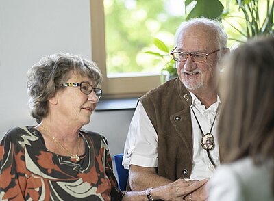 Ein älteres Paar sitzt zusammen in einem hellen Raum. Die Frau mit lockigem Haar trägt eine bunte Bluse und schaut den Mann lächelnd an. Der Mann hat ein freundliches Gesicht, trägt eine Weste und eine Kette. Hinter ihnen sind Pflanzen und Fenster zu sehen.