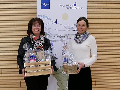 Zwei Frauen stehen nebeneinander und halten Geschenkkörbe in den Händen. Sie stehen vor einer Wand mit einer Karte und dem Logo "Allgäu". Die Frauen tragen Schals und lächeln, während sie in die Kamera schauen.