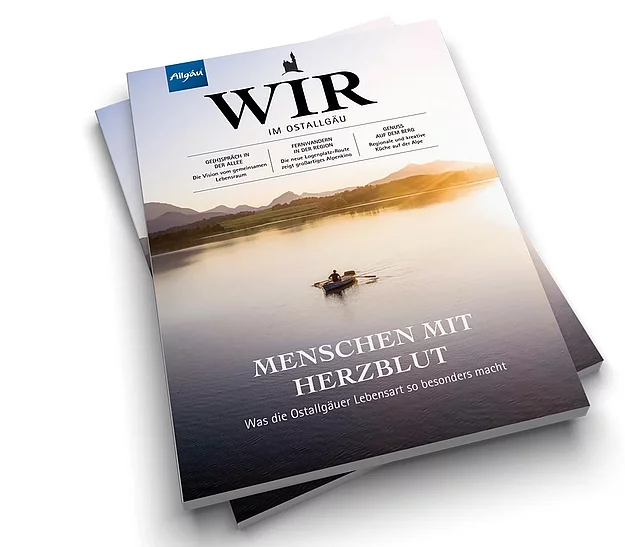 © Landkreis Ostallgäu Zwei Magazine liegen übereinander. Das obere zeigt den Titel "WIR" und den Untertitel "MENSCHEN MIT HERZ/BLUT". Im Hintergrund ist eine ruhige Wasseroberfläche und eine Person in einem Boot zu sehen, die auf einem Gewässer paddelt.