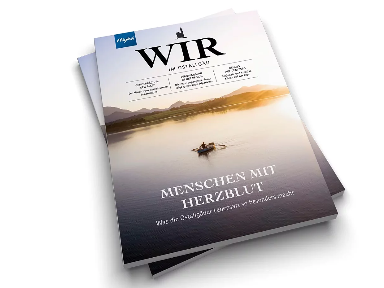 © Landkreis Ostallgäu Zwei Magazine liegen übereinander. Das obere zeigt den Titel "WIR" und den Untertitel "MENSCHEN MIT HERZ/BLUT". Im Hintergrund ist eine ruhige Wasseroberfläche und eine Person in einem Boot zu sehen, die auf einem Gewässer paddelt.