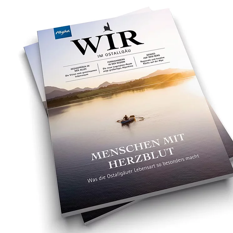 © Landkreis Ostallgäu Zwei Magazine liegen übereinander. Das obere zeigt den Titel "WIR" und den Untertitel "MENSCHEN MIT HERZ/BLUT". Im Hintergrund ist eine ruhige Wasseroberfläche und eine Person in einem Boot zu sehen, die auf einem Gewässer paddelt.