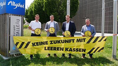 Vier Männer stehen vor einem Schild des Landratsamts Ostallgäu. Sie halten große Uhren in den Händen und stehen vor einem gelben Banner mit der Aufschrift „KEINE ZUKUNFT MIT LEEREN KASSEN“. Im Hintergrund sind Bäume und ein modernes Gebäude sichtbar.